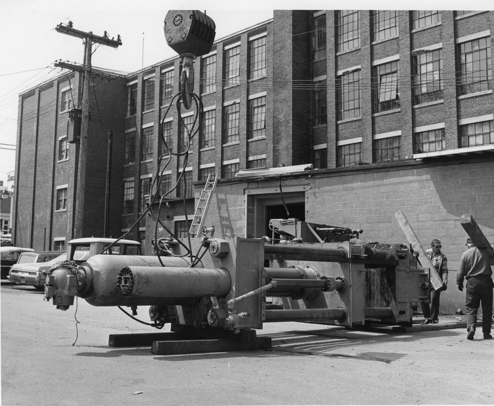 A crane hook lifts a huge hydraulic press off timber blocks in the street beside the brick factory